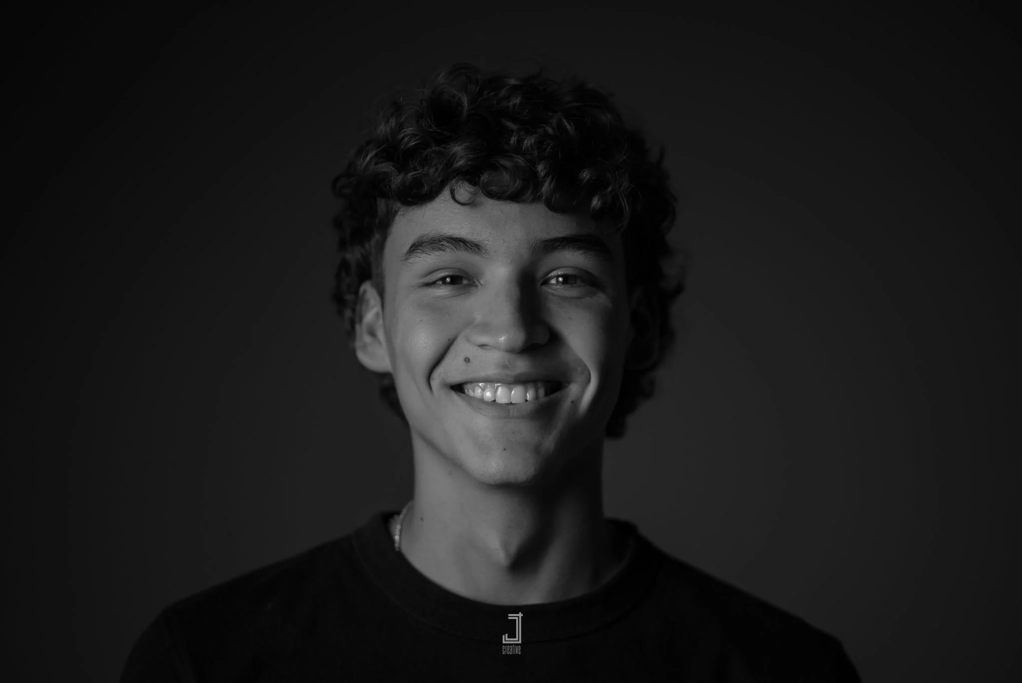 Black and white portrait of a young person with curly hair, smiling, wearing a dark shirt against a dark background.