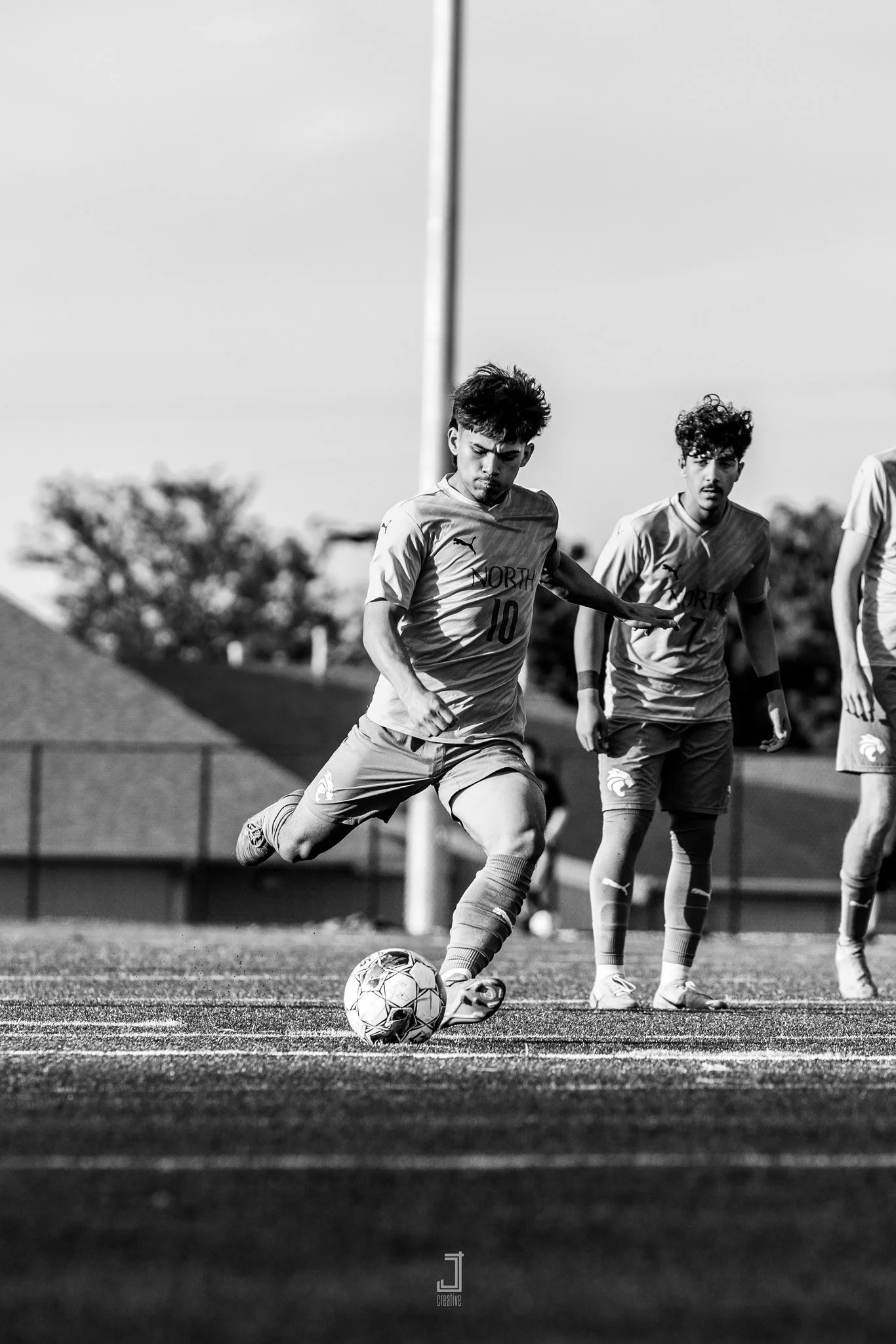 Black and white photo of young male soccer players on field, one about to kick soccer ball, wearing North Shore jerseys, others watching, trees and fence in background.
