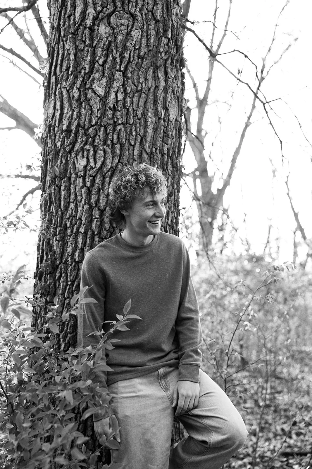 A young man with curly hair smiling and leaning against a large tree trunk in a forest, wearing a sweatshirt and jeans, black and white photograph.