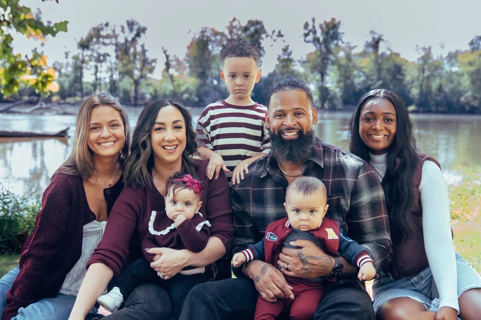 A diverse family of eight posing by a river with trees in the background, smiling and sitting on the grass.