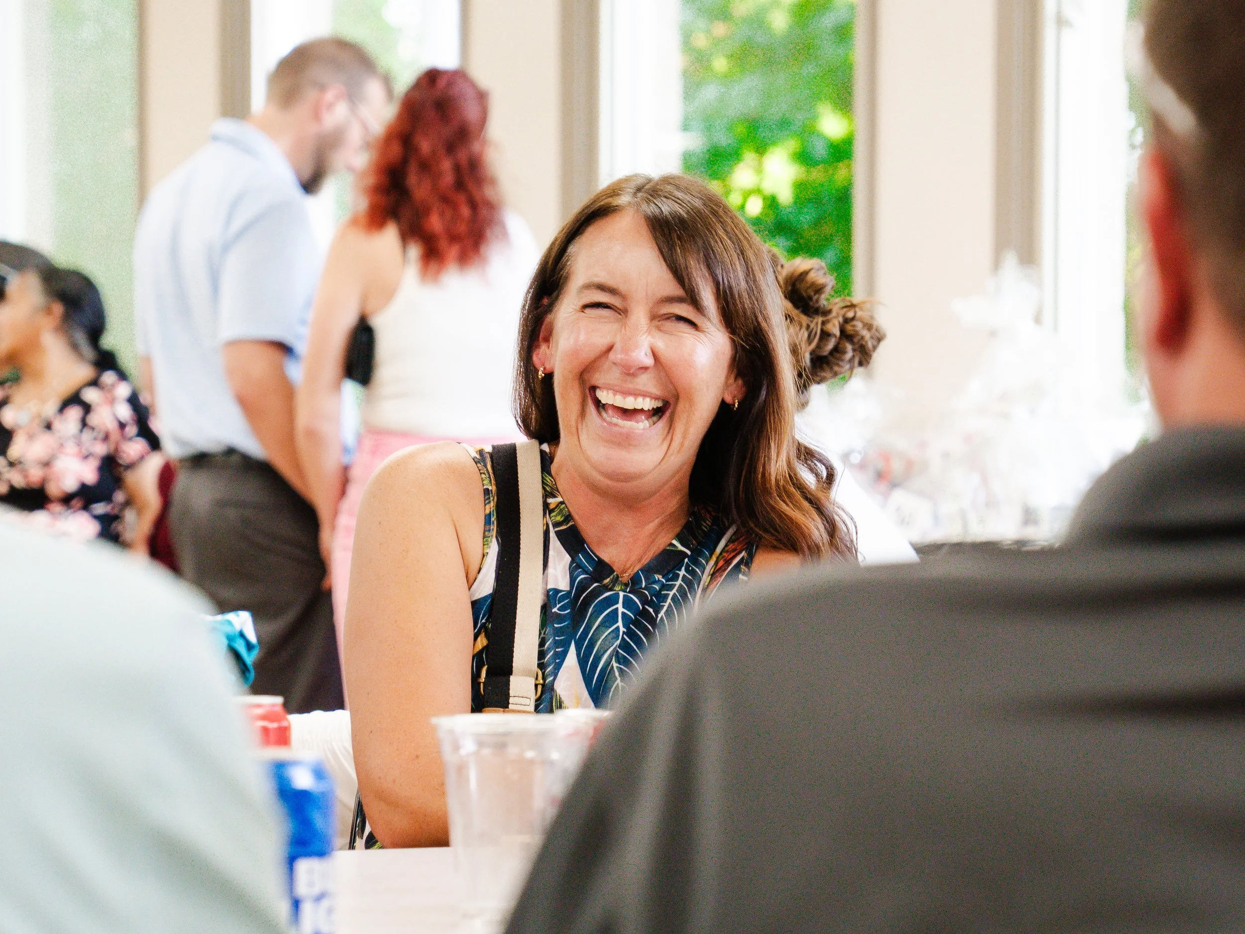 A woman laughing with others at a social gathering near a window with green trees outside.
