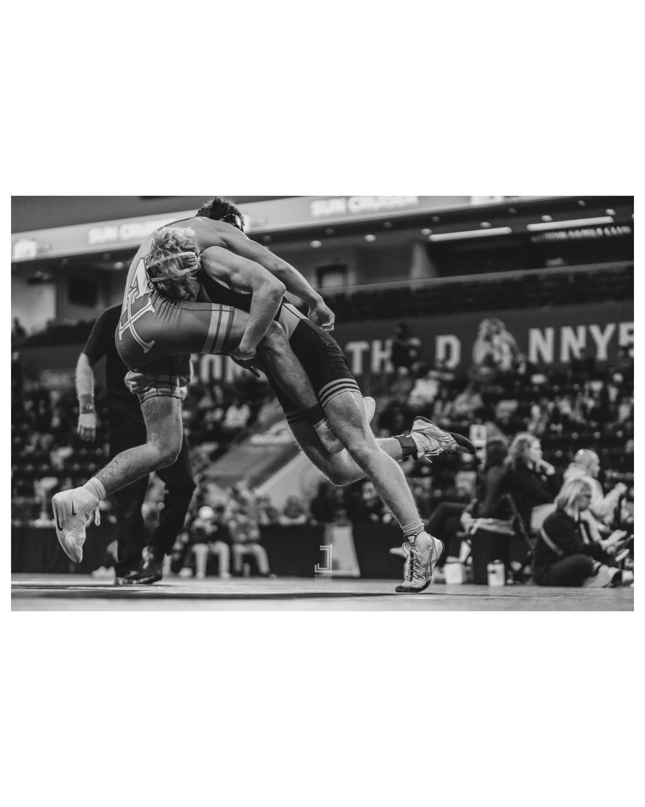 Black and white photo of two wrestlers engaged in a match inside a gymnasium with an audience watching in the background.