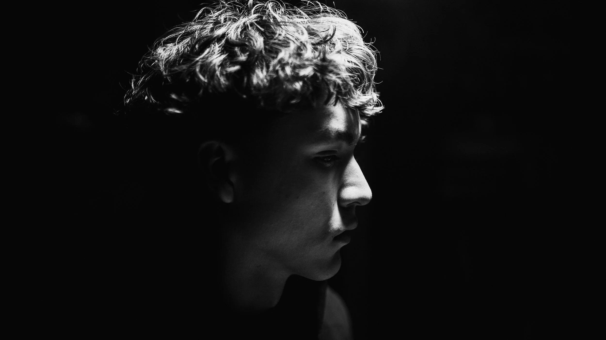Side profile of a young person with curly hair, illuminated from the side, against a dark background.