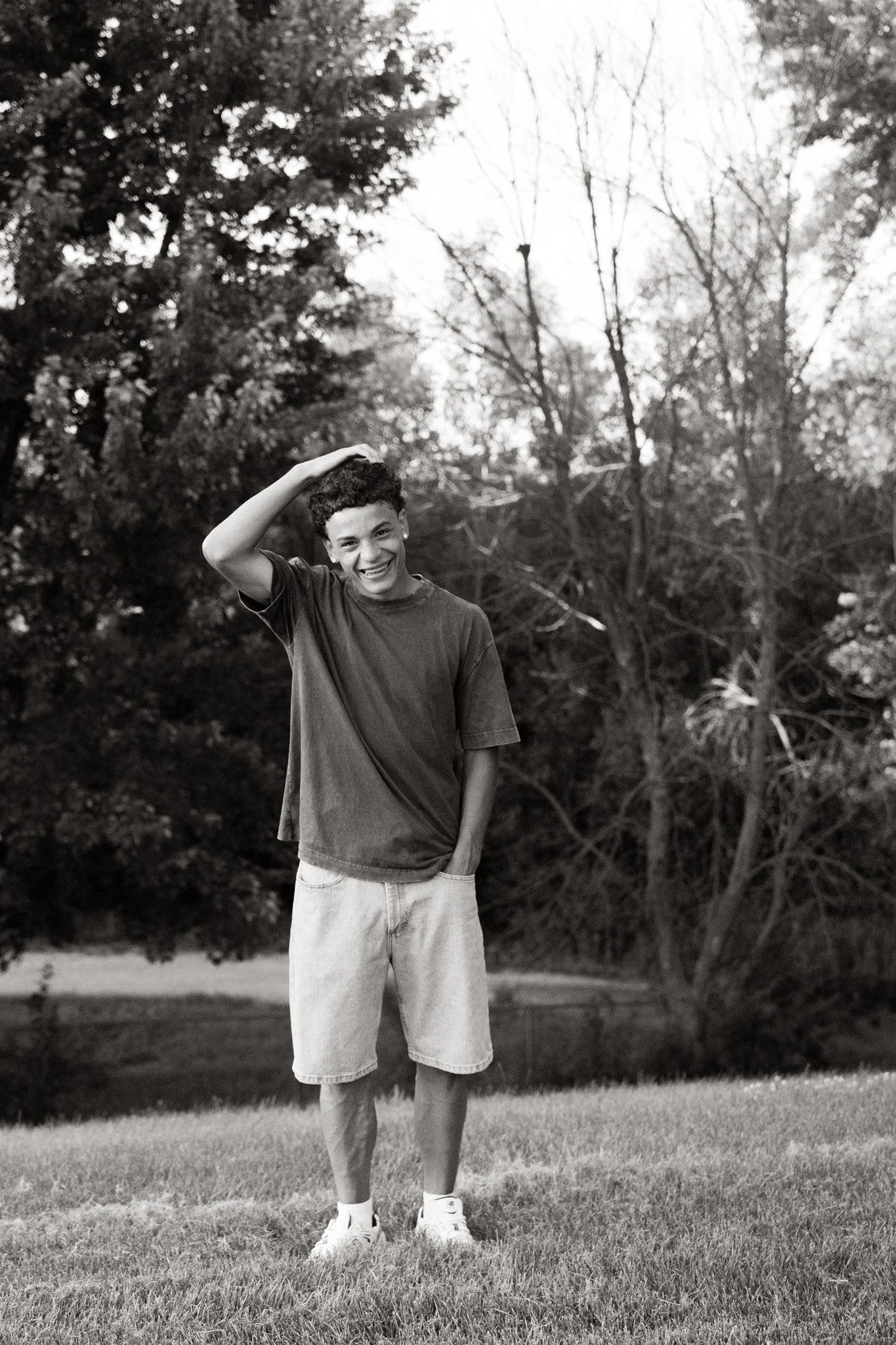 A smiling young man standing outdoors on grass, wearing a t-shirt, shorts, and sneakers, with trees in the background.