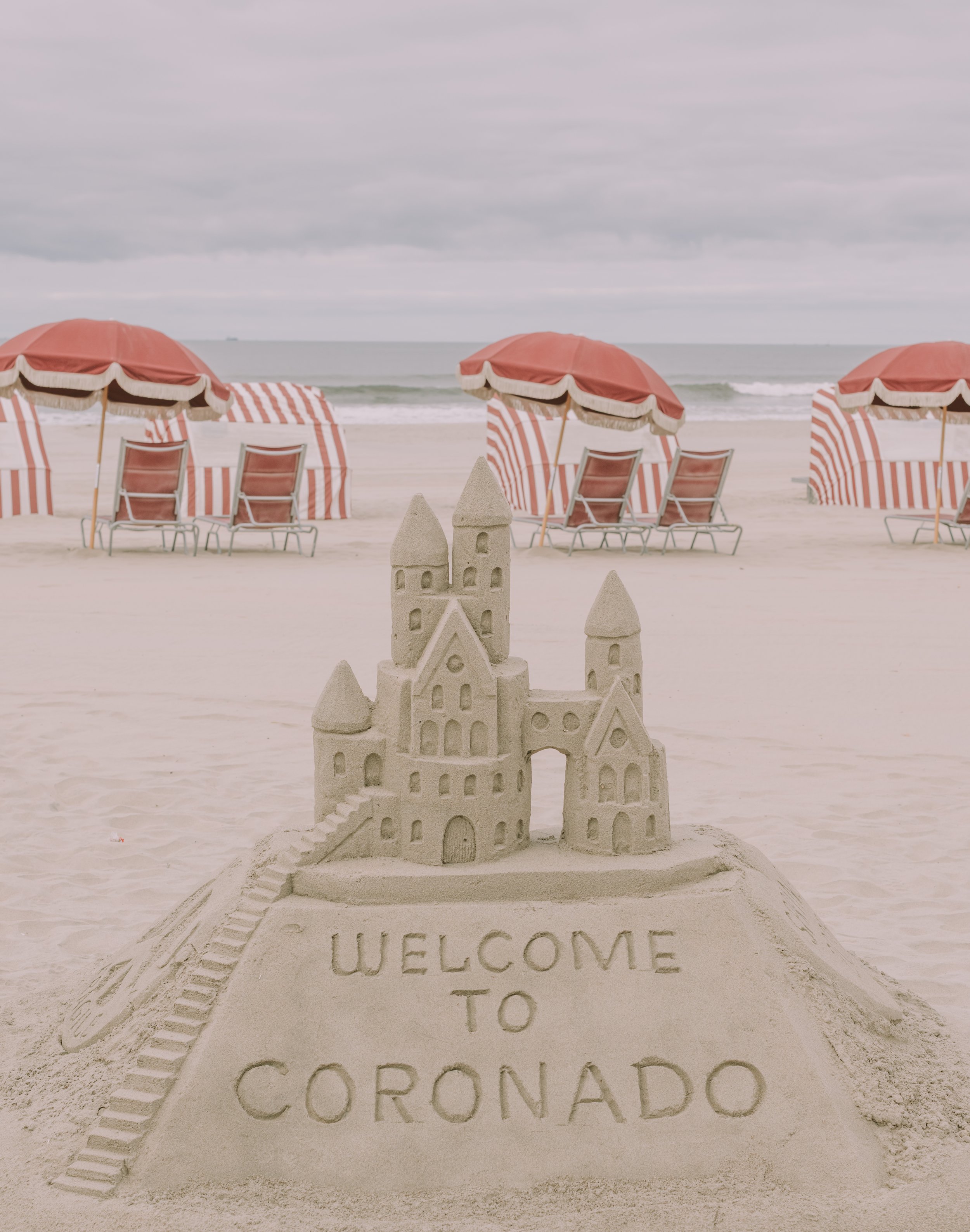 Sandcastle on the beach with the words 'Welcome to Coorado' inscribed in the sand, surrounded by umbrellas and chairs overlooking the ocean.