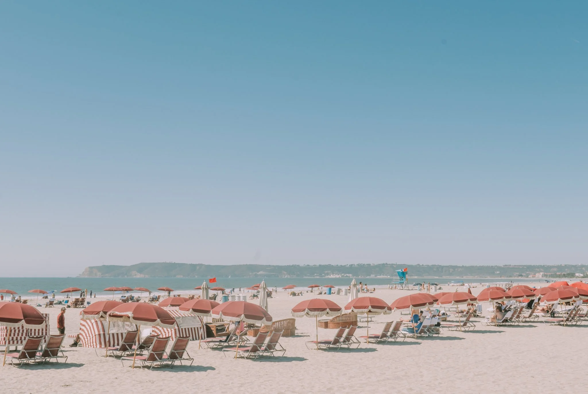 Beach scene with red umbrellas, lounge chairs, people walking and relaxing, lifeguard tower, ocean, cliffs in the background, and a clear blue sky.