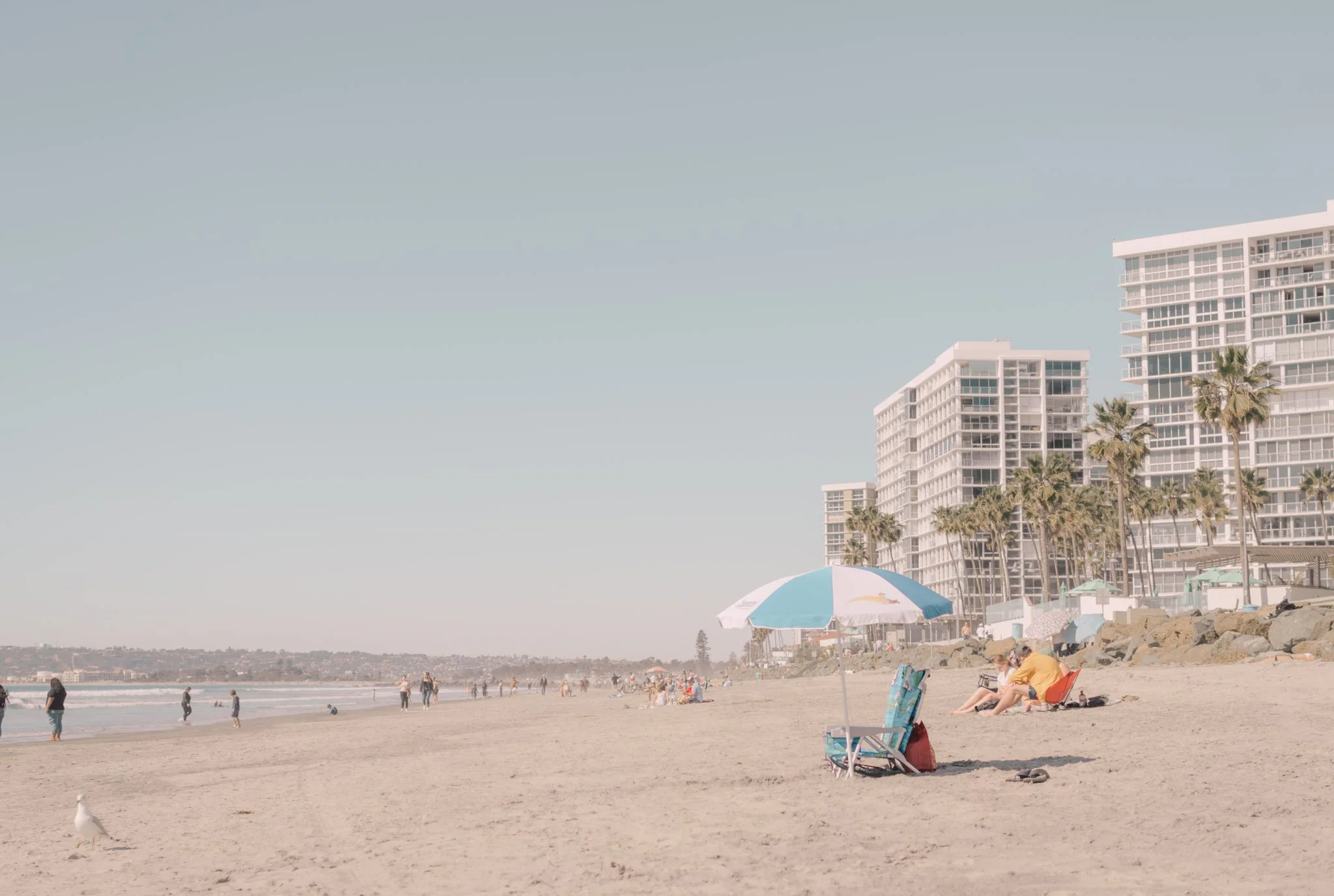 People relaxing and walking on a sandy beach with umbrellas, high-rise buildings, and palm trees in the background on a sunny day.