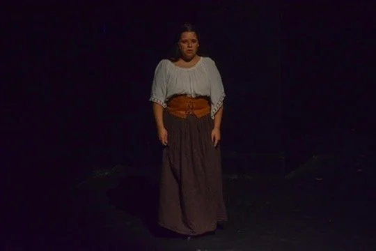 A woman standing alone on a dark stage, wearing a white blouse and a long dark skirt.