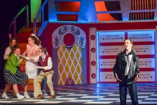 A theatrical stage with five actors in vintage clothing performing. Four actresses are on the left, holding hands and smiling; one actor is on the right, standing alone. The background has colorful stairs and a decorative set with a large opening.