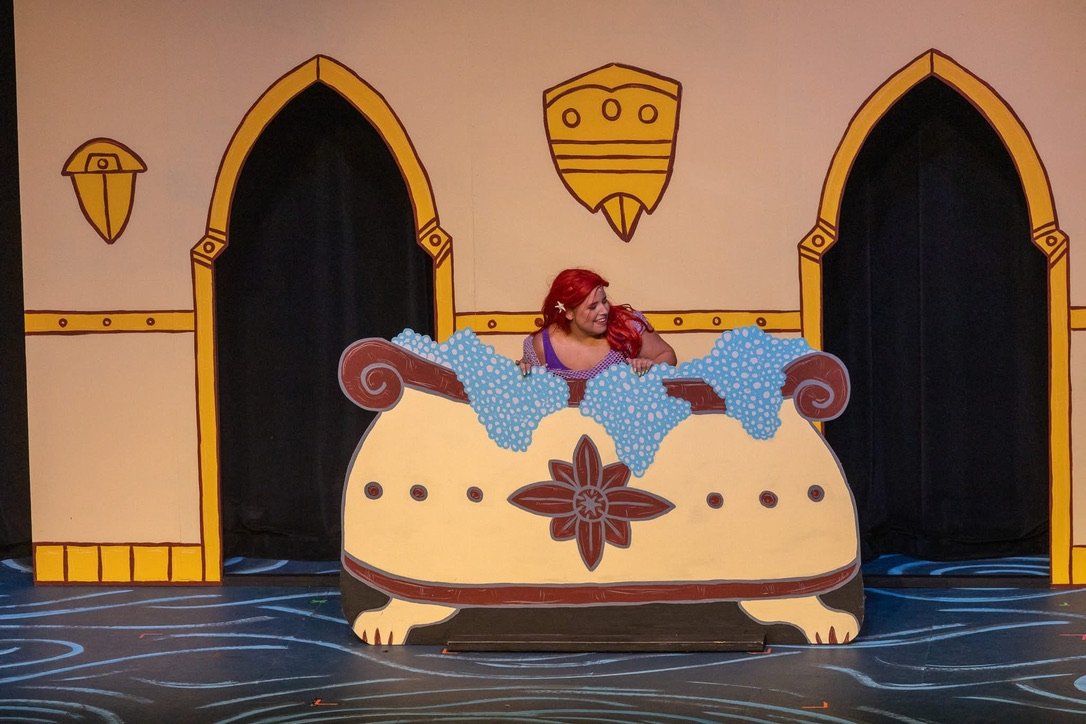 A person with red hair, dressed as Ariel from The Little Mermaid, is leaning out of a cardboard bathtub decorated with sea-themed designs, in front of a painted backdrop resembling a castle interior with arched doorways and medieval decoration.