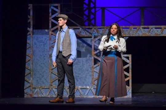 Two actors on stage in costume, with a metal scaffold and blue lighting in the background.