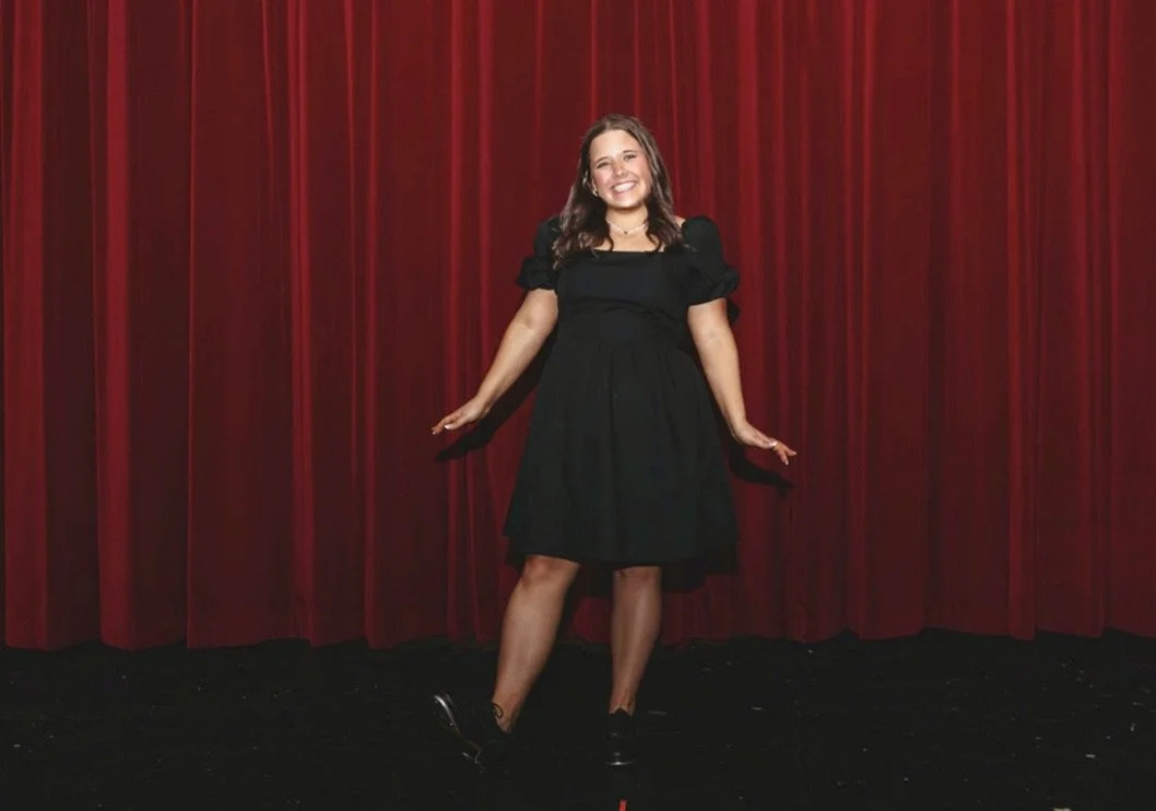 A young woman with brown hair and a big smile standing in front of red theater curtains, wearing a black dress and black shoes.
