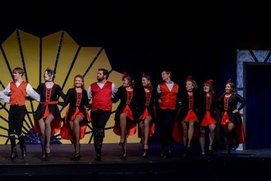 Group of performers on stage dressed in colorful costumes, posing in a dance formation with a geometric backdrop.