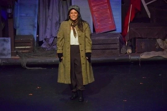 Woman standing on stage with colorful set pieces in the background.