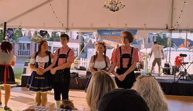 Group of young people in traditional Bavarian attire performing on a stage at an outdoor event under a white tent with string lights.