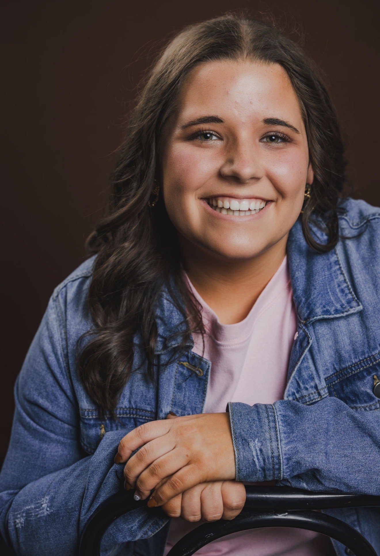 Close-up of a smiling woman with brown hair, blue eyes, wearing a denim jacket over a light pink shirt, sitting with her hands resting on a black railing.