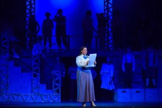 A woman is on stage reading a paper in front of a group of people, with blue lighting and stage scaffolding visible.
