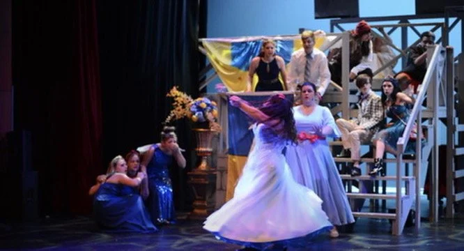 The scene shows a theatrical performance with a group of children on stage. Some are sitting on the stairs, while others are standing or dancing. The stage is decorated with colorful banners and flowers.