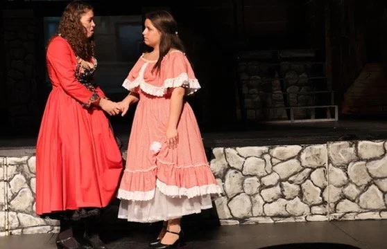 Two women dressed in period costumes holding hands on a stage with a stone wall backdrop.