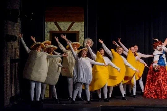A group of people dressed in costumes performing on stage, some in yellow dresses and others in old-fashioned attire, possibly participating in a theatrical production.