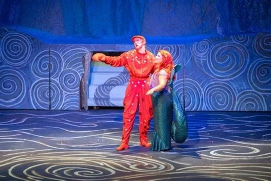 Two performers dressed in colorful costumes on a stage set with ocean-themed background and floor patterns, acting in a theatrical production.