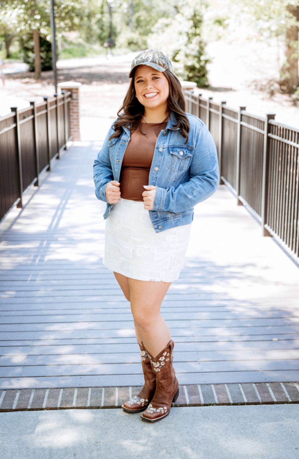 A young woman stands outdoors on a wooden bridge, smiling at the camera. She is wearing a denim jacket, brown top, white skirt, cowboy boots, and a cap, with trees and sunlight in the background.