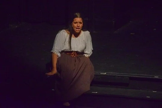 A woman sitting on a dark stage, appearing to sing or speak, with dark background and stage lighting.