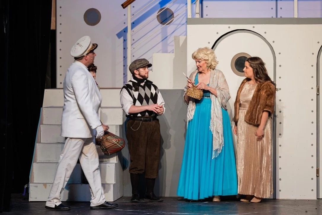 Five actors on a stage performing in a play, with a backdrop resembling a nautical or maritime theme. They are dressed in costumes, including a captain's uniform, vintage clothing, and period costumes.