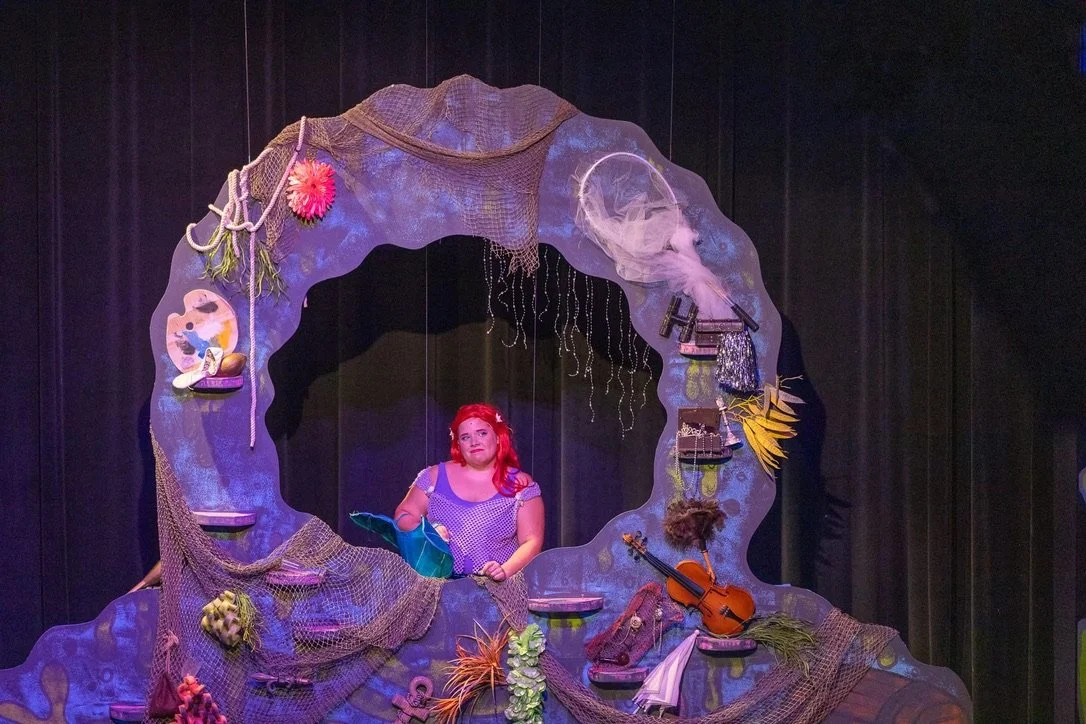 A woman with red hair standing behind a decorative window frame with various nautical decorations and a violin. Dark curtains in the background.