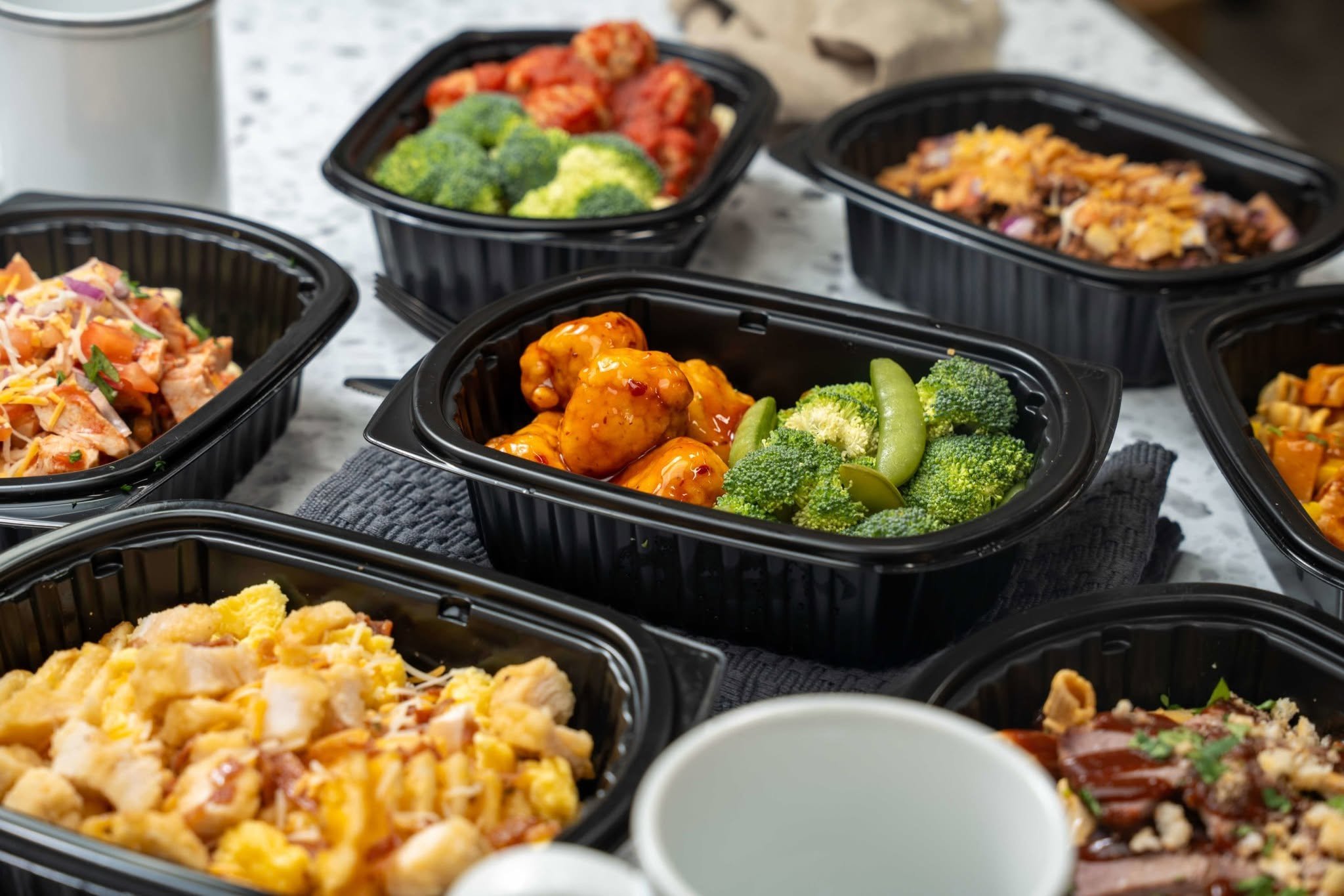 Various prepared dishes in black plastic containers, including broccoli, red peppers, shredded cheese, fried chicken, and pasta with sauce on a marble countertop.