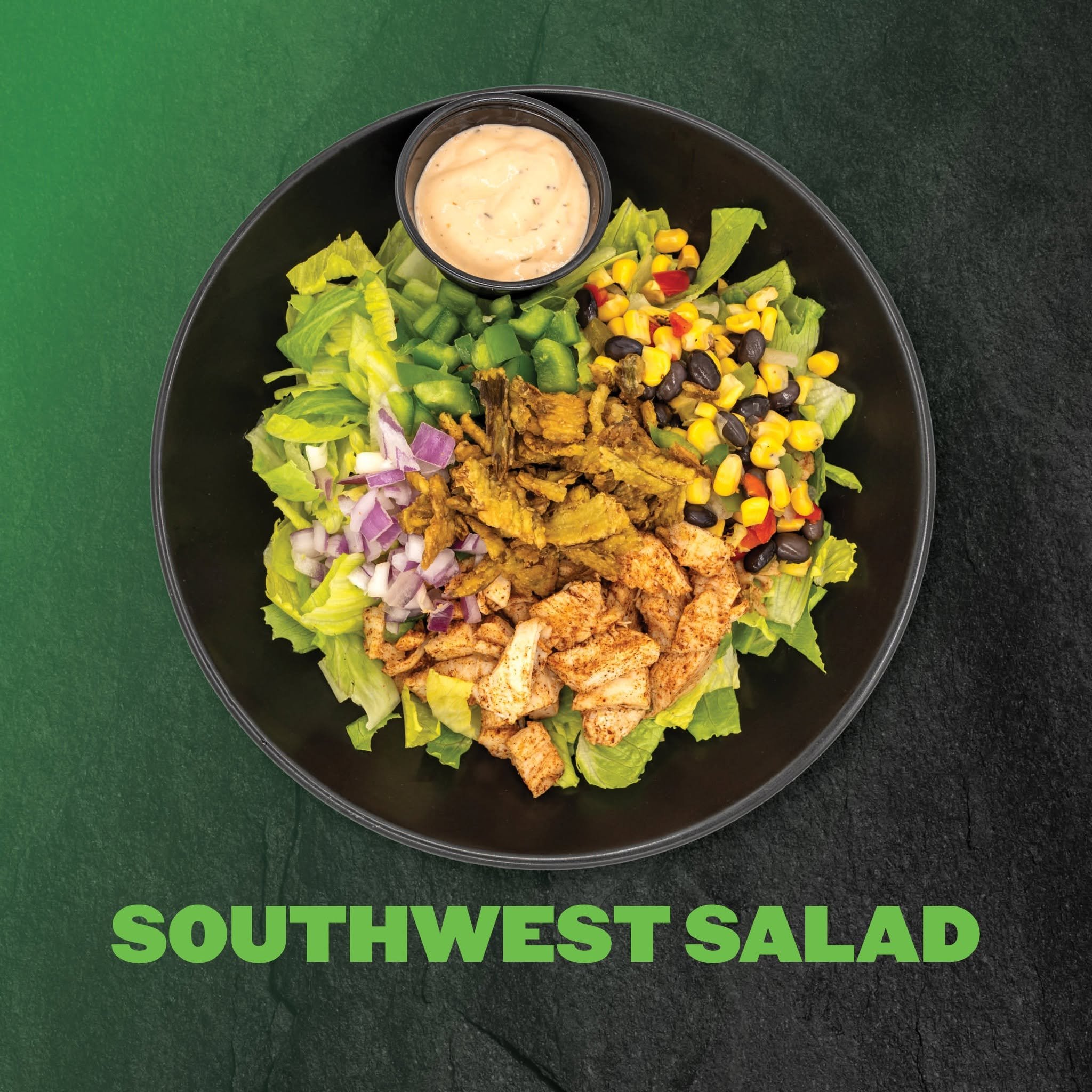 A bowl of Southwest salad with diced lettuce, corn, black beans, chopped onions, and grilled chicken slices, topped with a creamy dressing on a black plate against a green background.