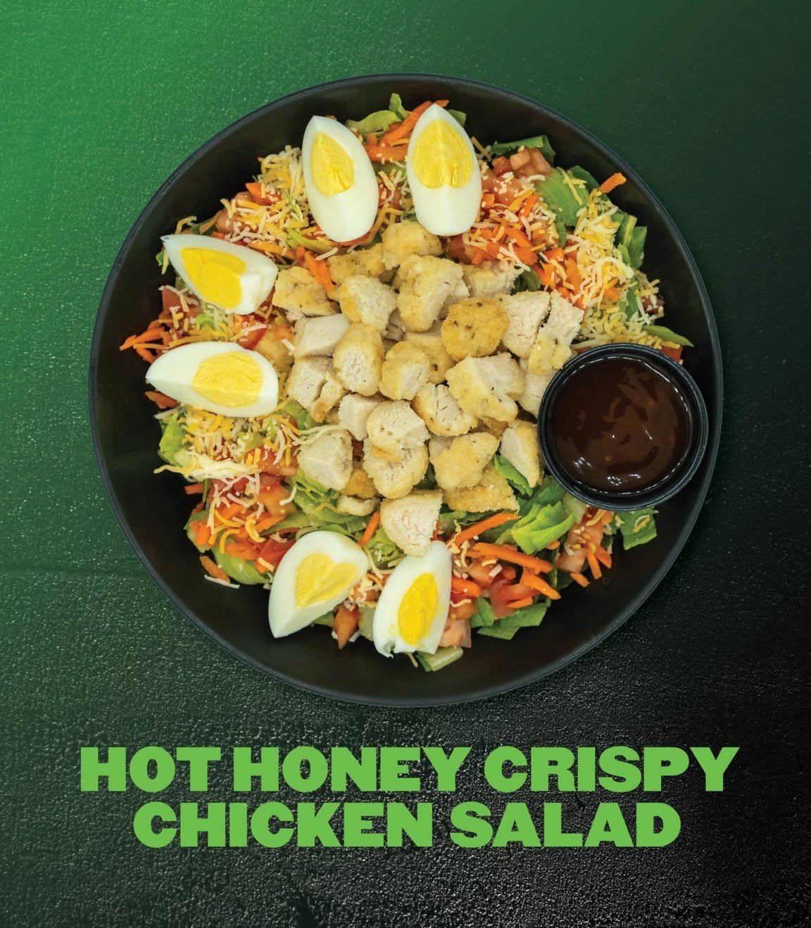 Bowl of salad with shredded lettuce, carrots, diced tomatoes, shredded cheese, sliced boiled eggs, and crispy chicken pieces with a small cup of dressing on a green surface. Text overlay says 'Hot Honey Crispy Chicken Salad'.