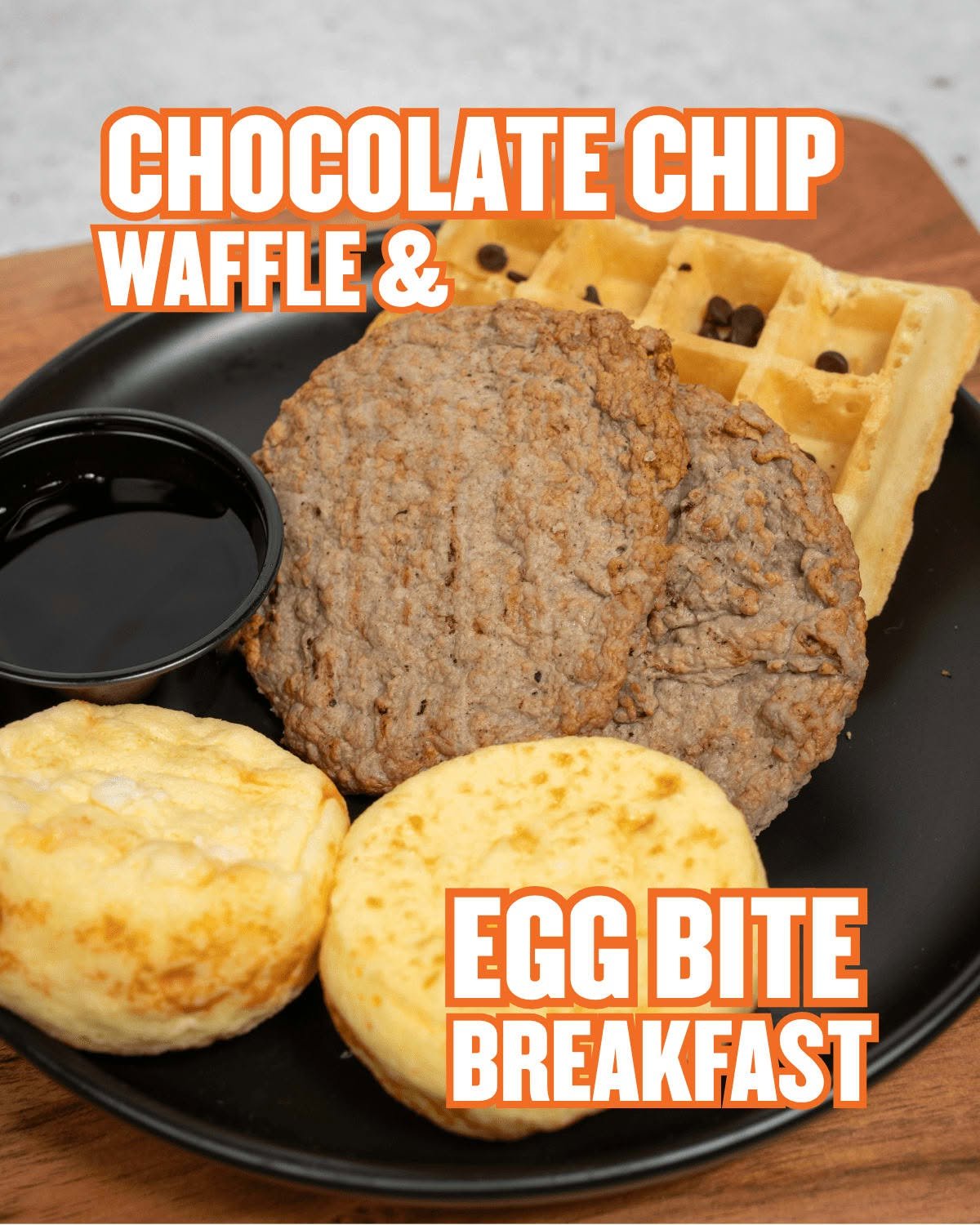 A plate with two breakfast biscuits, a small cup of syrup, a chocolate chip waffle, and a sausage patty. Text overlay reads 'Chocolate Chip Waffle & Egg Bite Breakfast'.
