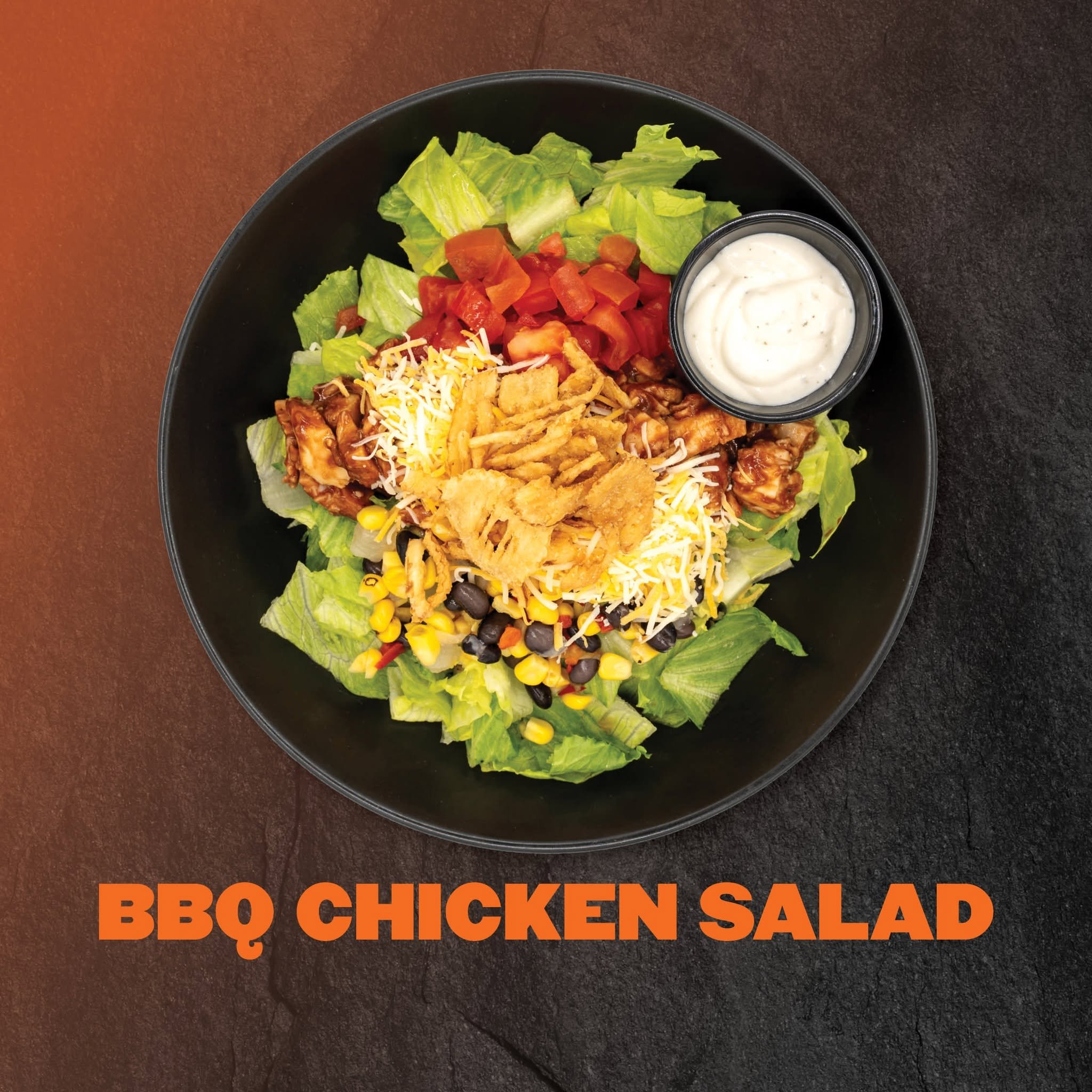 Black round plate with lettuce, chopped tomatoes, shredded cheese, fried onion strings, grilled chicken pieces, black beans, corn, and a side cup of creamy salad dressing, with a part of a branded orange and black sign with the words "BBQ CHICKEN SAL
