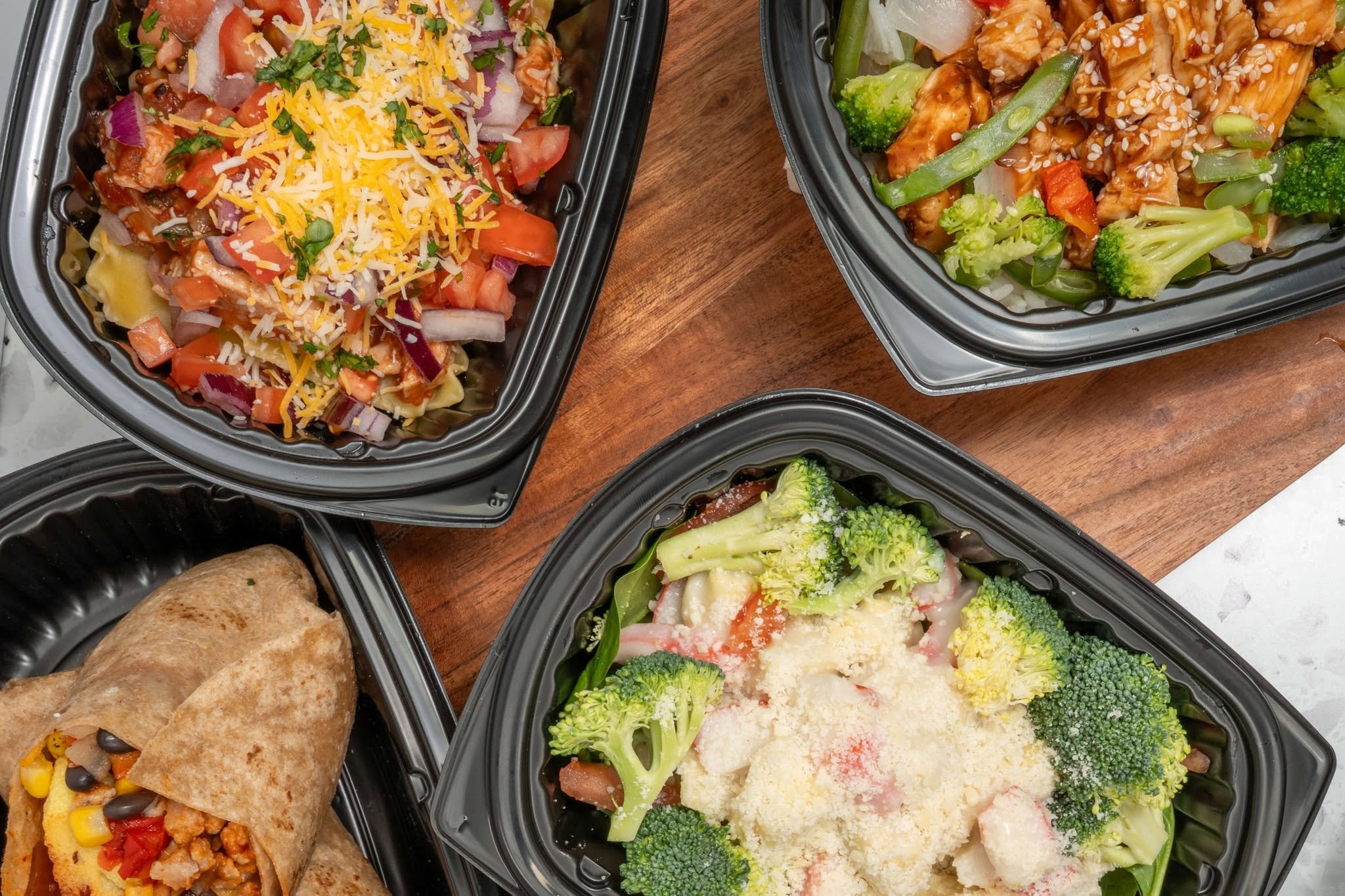 Multiple takeout dishes with salads, broccoli, and a burrito, served in black plastic containers on a wooden table.