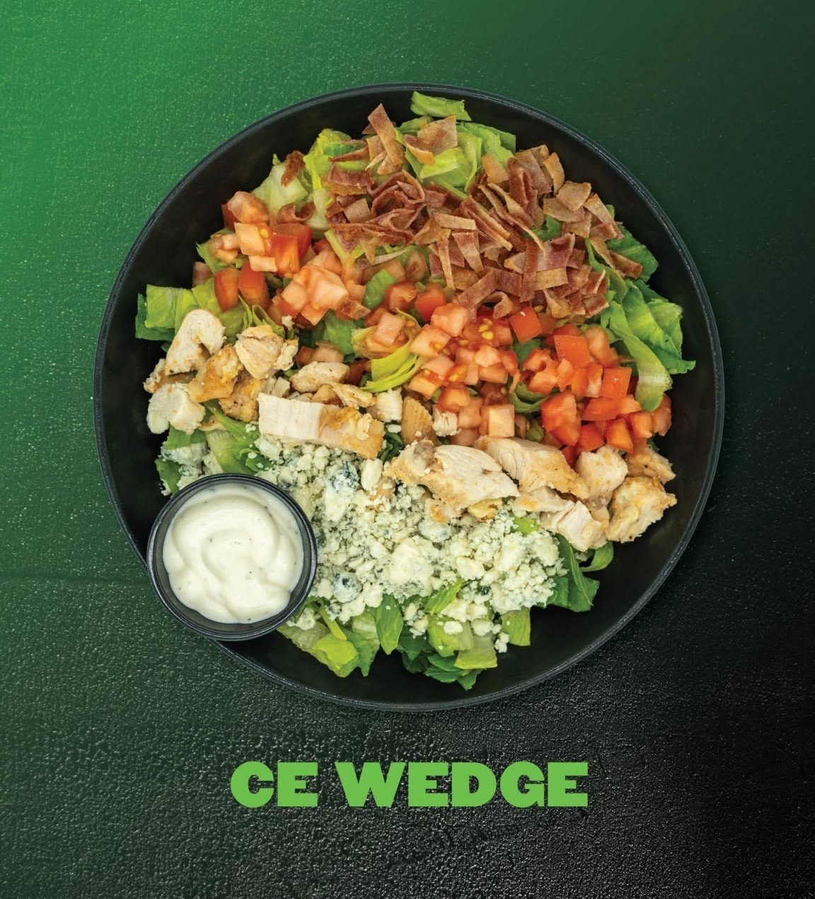 Salad in a black bowl with lettuce, diced tomatoes, diced cucumbers, shredded chicken, blue cheese crumbles, bacon bits, and a side container of salad dressing. Green textured background. The text 'CE WEDGE' below the bowl.