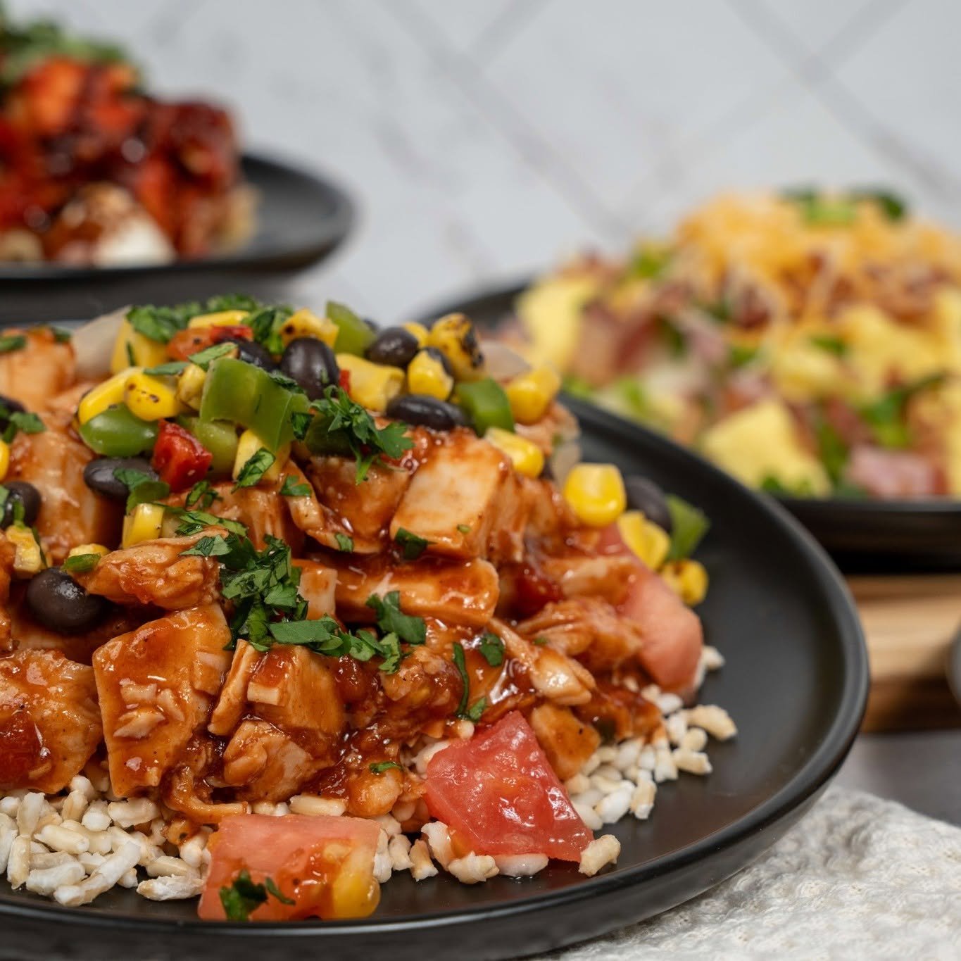 Close-up of chicken and vegetable stir-fry topped with chopped herbs, served on a bed of rice. In the background, two other plates with similar dishes are slightly out of focus.