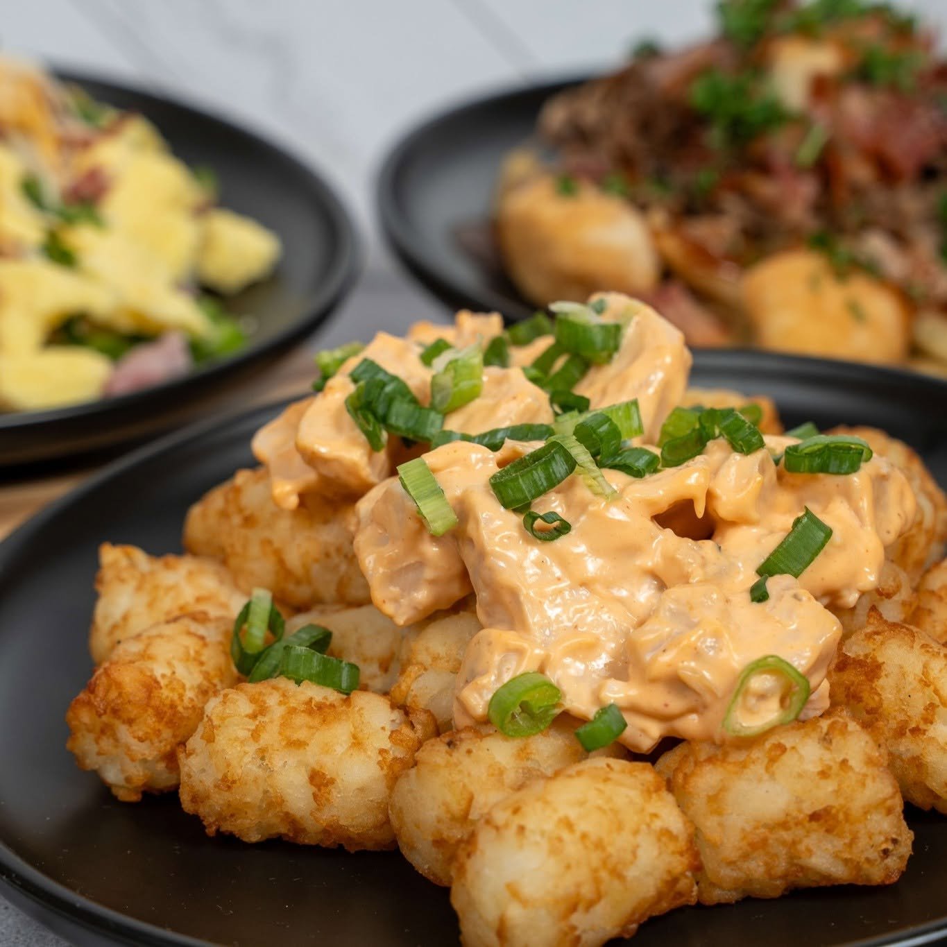 Fried fish topped with creamy sauce and chopped green onions, with two other dishes in the background on black plates.