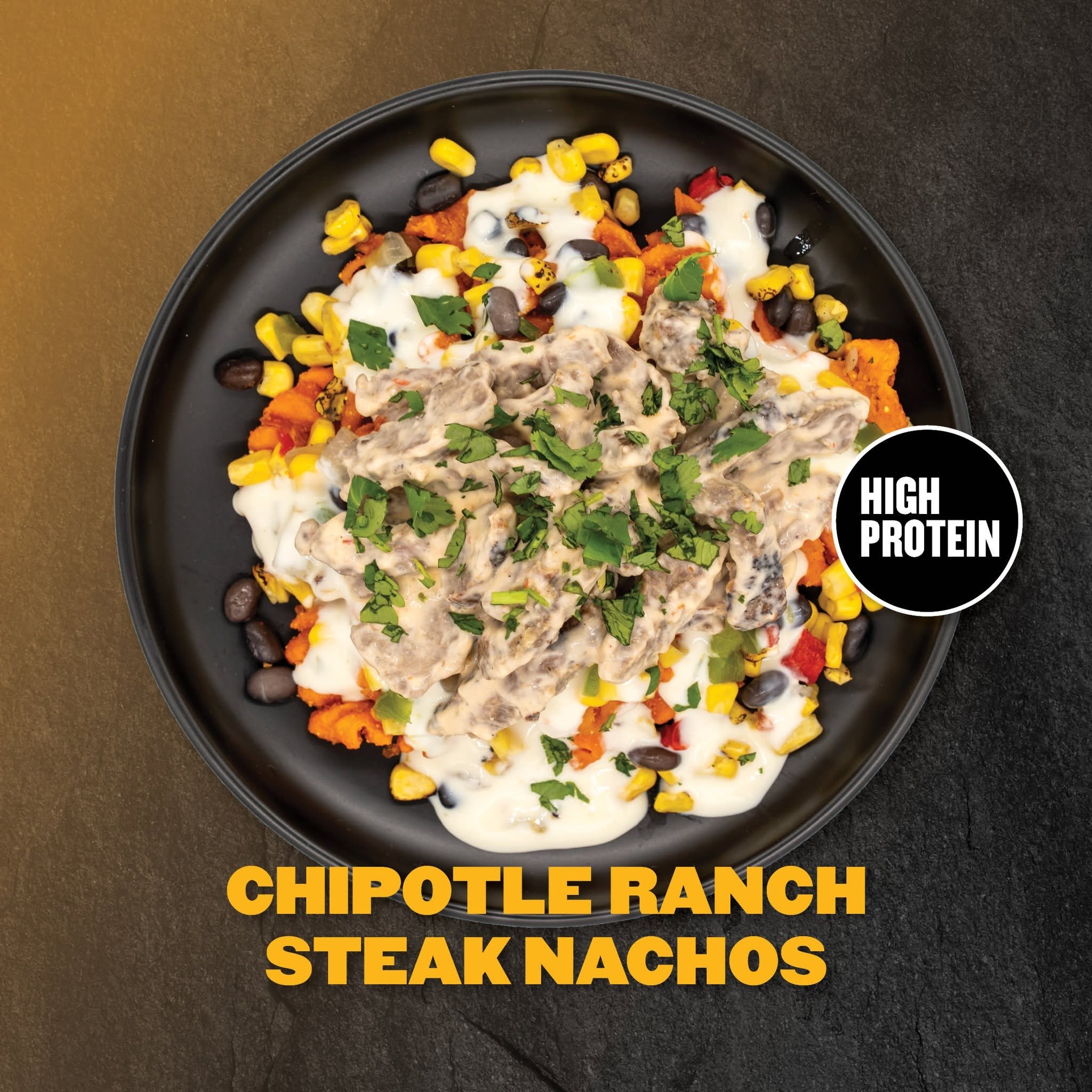 A black oval plate contains a Mexican-style dish with meat, corn, black beans, cheese, sour cream, red peppers, and chopped cilantro on a dark surface. Text overlay says 'HIGH PROTEIN CHIPOTLE RANCH STEAK NACHOS'.