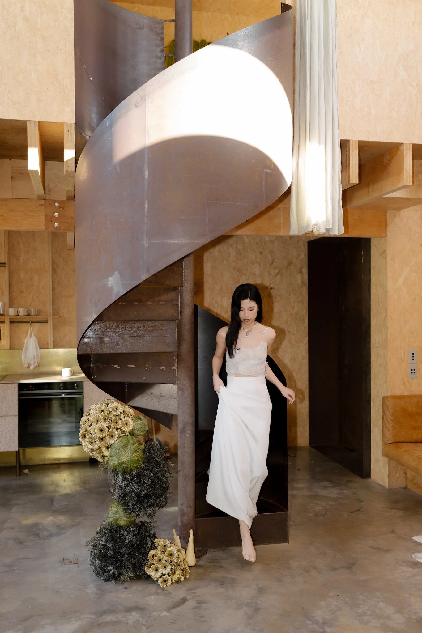 A woman in a white dress standing on one foot under a metal spiral staircase in a room with beige walls and a kitchen in the background. There are flower arrangements on the floor near her.