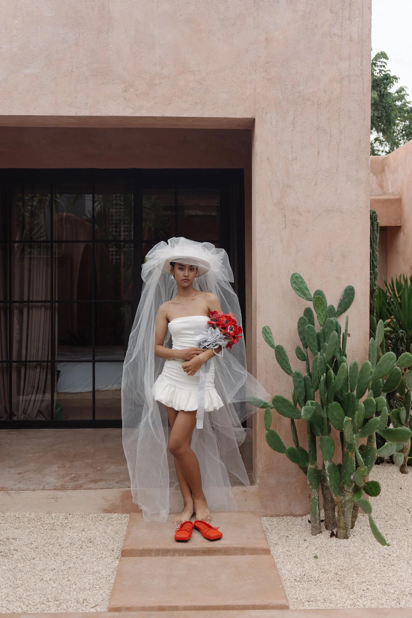 A woman standing in front of a pink wall with a cactus plant to her right, wearing a white dress, a large white hat with tulle, holding red flowers, and red shoes on the ground.