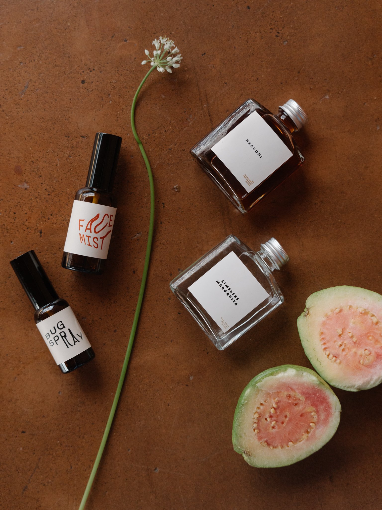 A flat lay of three small perfume bottles labeled 'Face Mist', 'Bug Spray', and 'Negroni', with a small white flower, a sliced green fig, and a green stem on a brown textured surface.