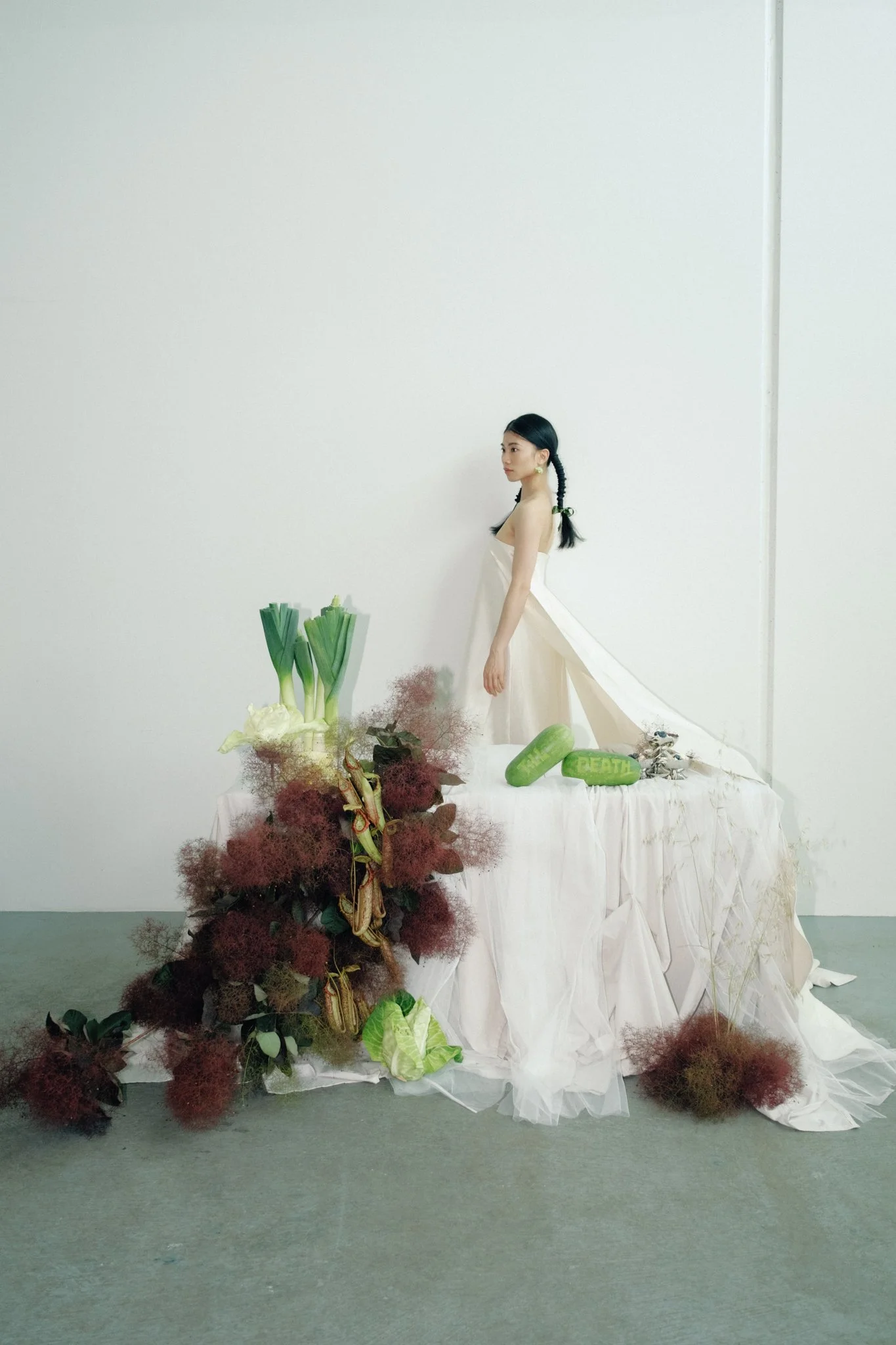 A young woman in a white dress with long black hair styled in braids, standing beside a table filled with flowers and plants, against a plain white wall.