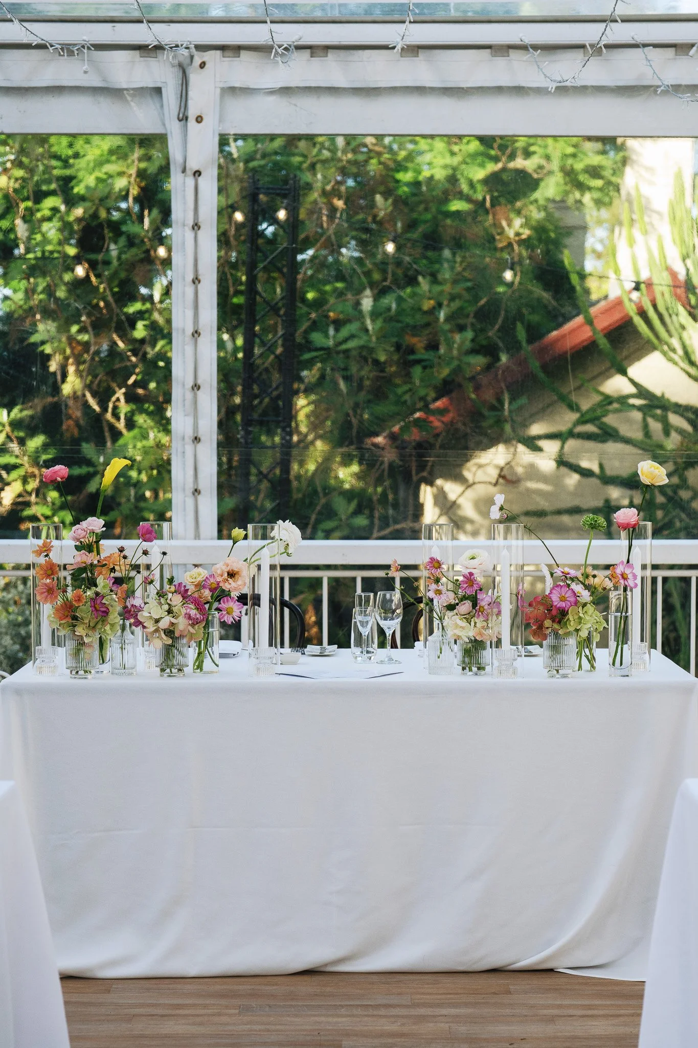 A banquet table covered with a white tablecloth, decorated with vases of pink, white, and peach flowers, and set with glasses and plates, situated near a window with a greenhouse view of greenery and string lights outside.