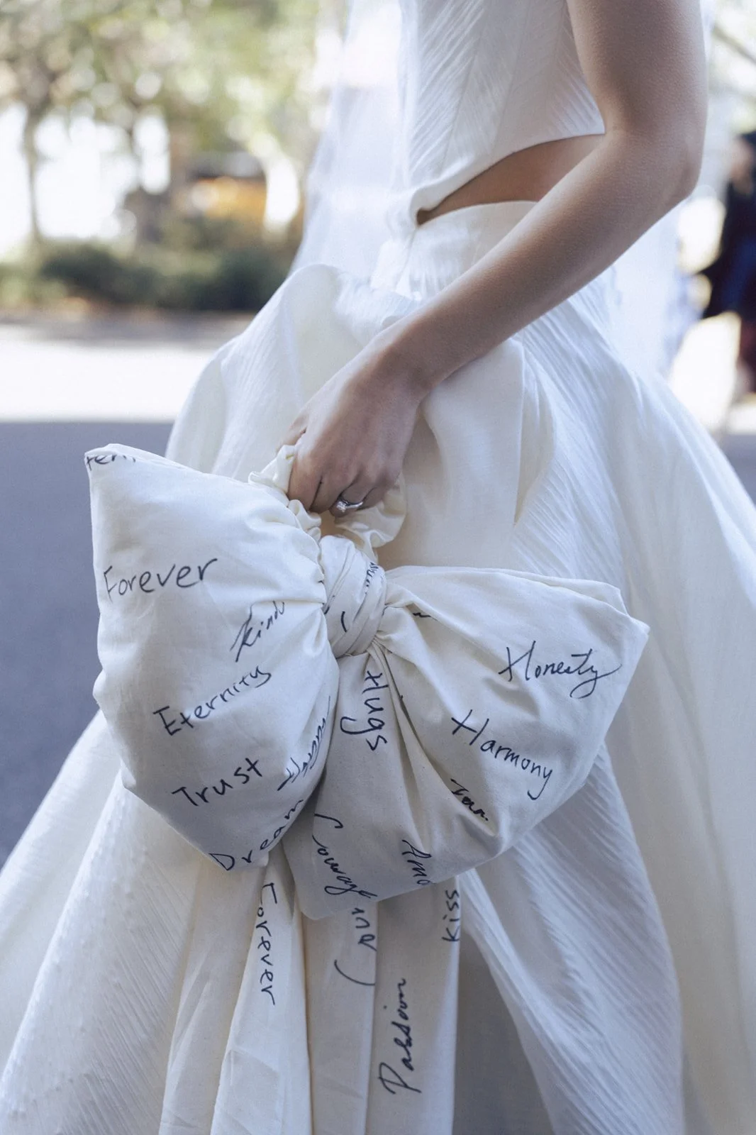 A woman in a white dress holding a large white fabric bundle with handwritten words like 'Forever,' 'Trust,' 'Happiness,' 'Harmony,' and 'Love,' written in black ink.
