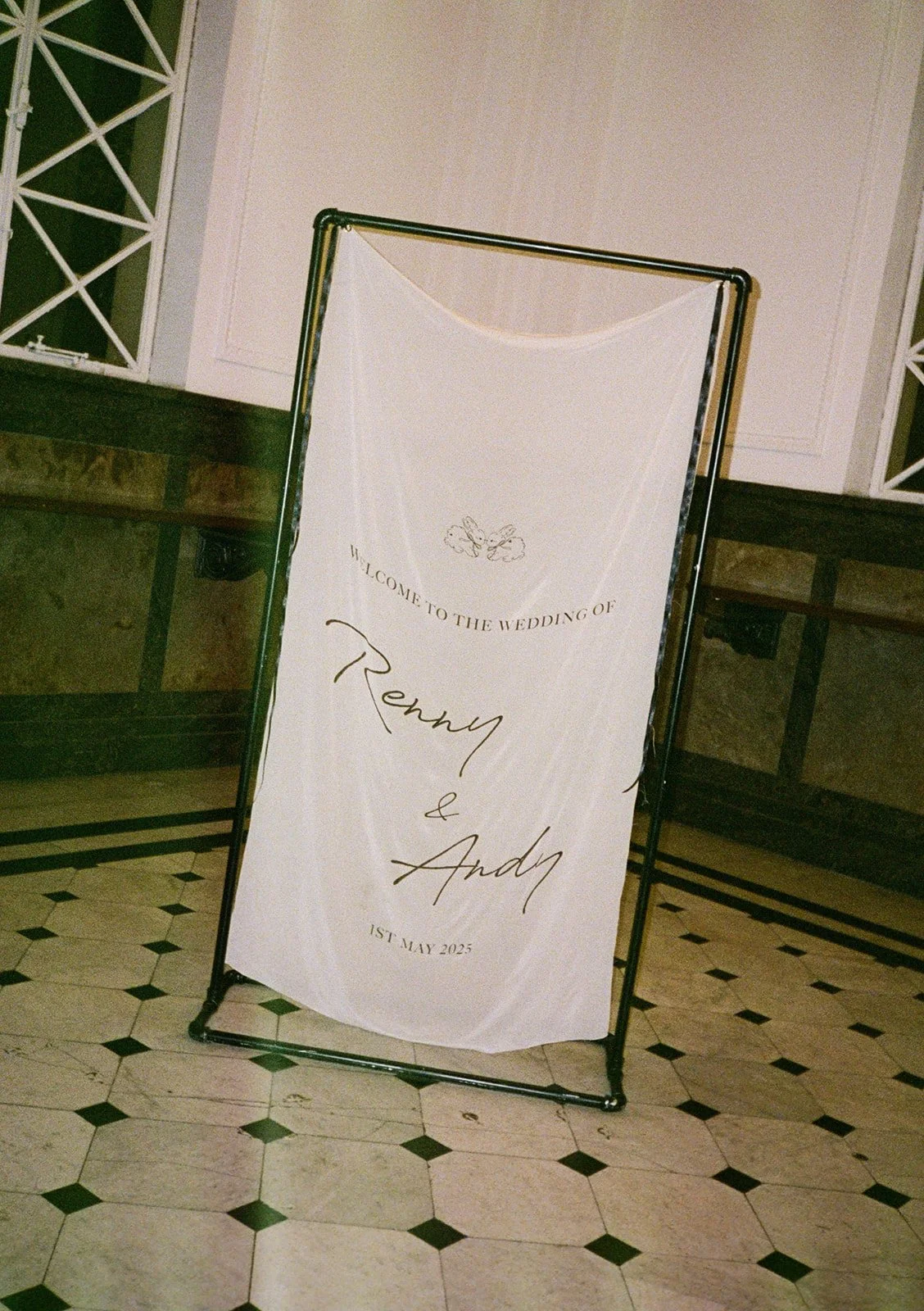 Wedding welcome sign for Ronny and Andy, dated May 1, 2025, with a white background, black text, and decorative bows at the top, placed on a black metal stand in an indoor setting.