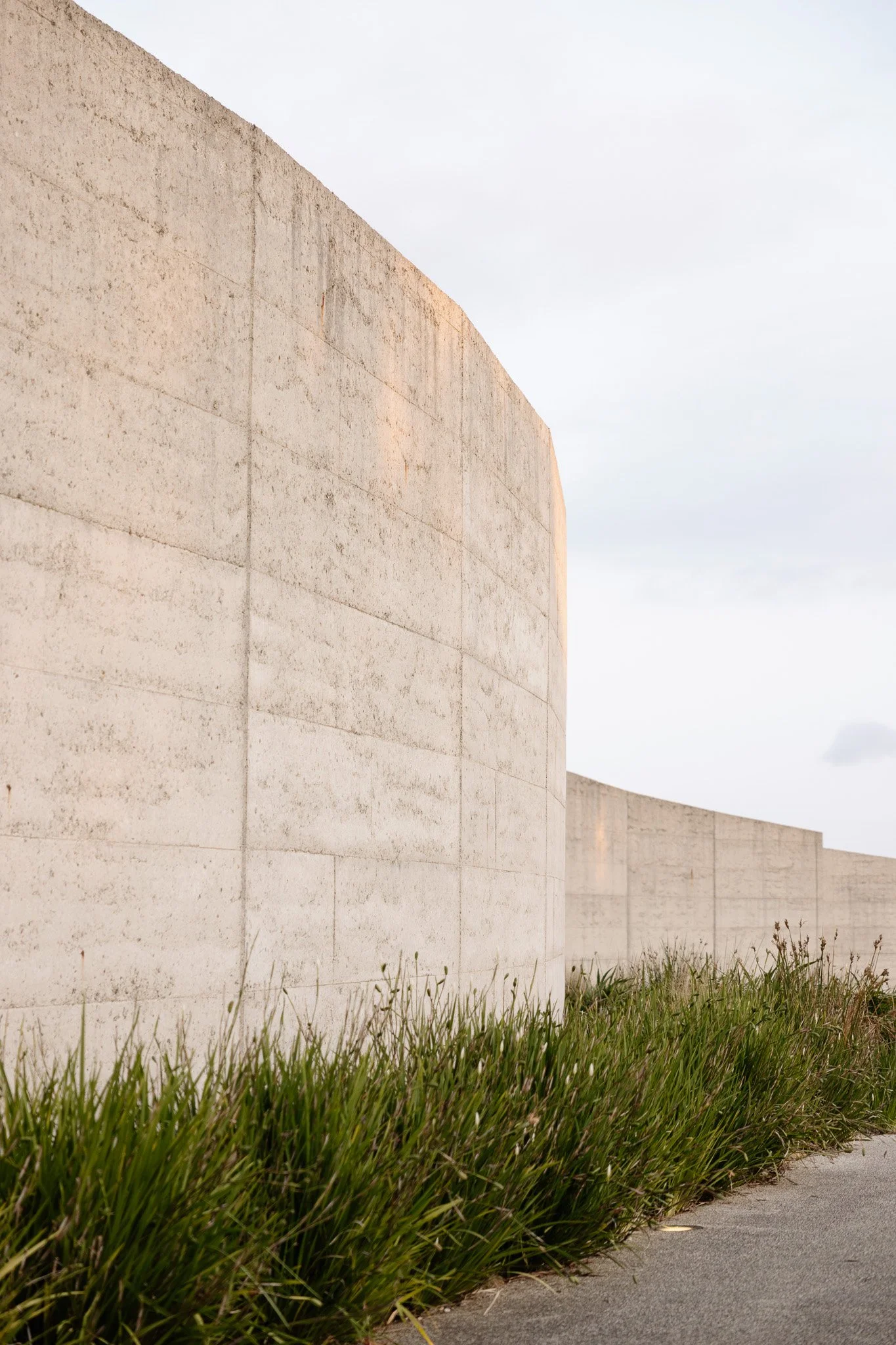 Close-up view of a large light-colored stone wall with a cloudy sky in the background. Green grass grows at the base of the wall along the sidewalk.