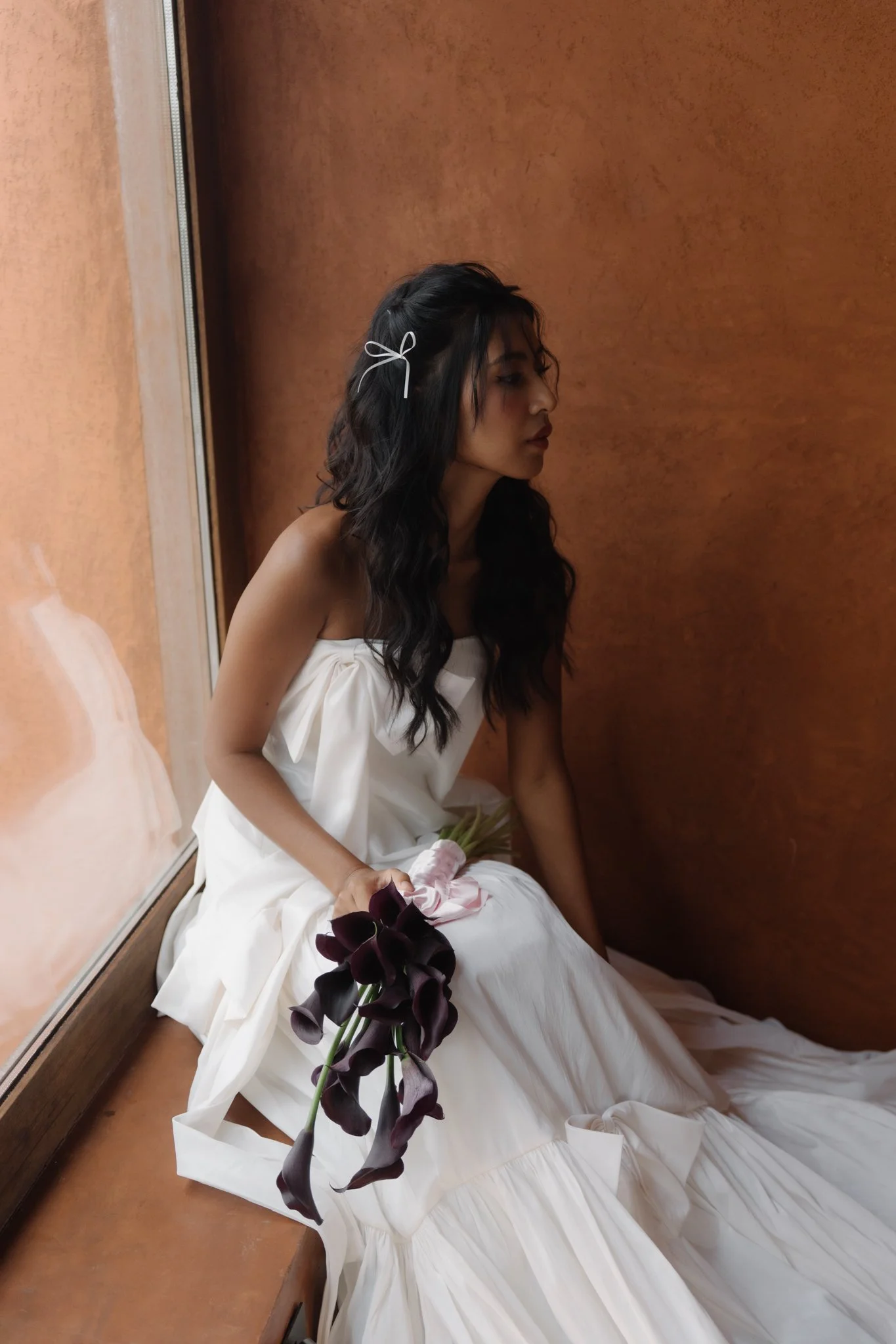 A woman with long dark hair, wearing a white dress, sitting on a wooden ledge next to a mirror, holding dark purple flowers, with her eyes closed and her head slightly tilted.