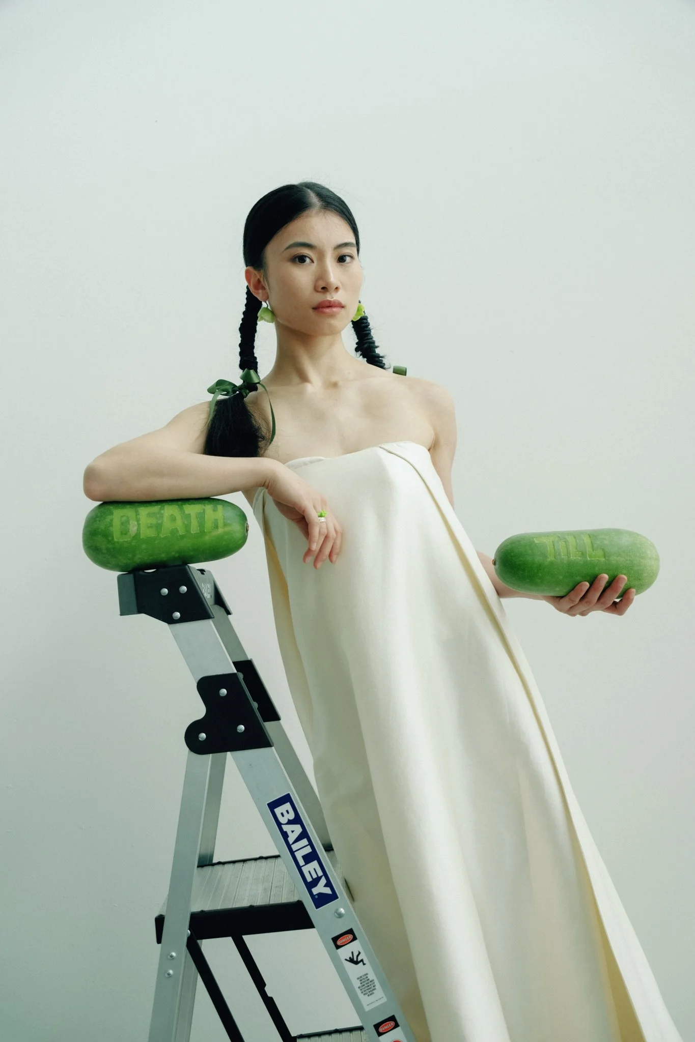 A woman with black hair styled in two braided pigtails tied with green ribbons. She is wearing a cream-colored dress and standing next to a step ladder holding two green watermelons, one labeled 'DEATH' and the other 'TILL'.