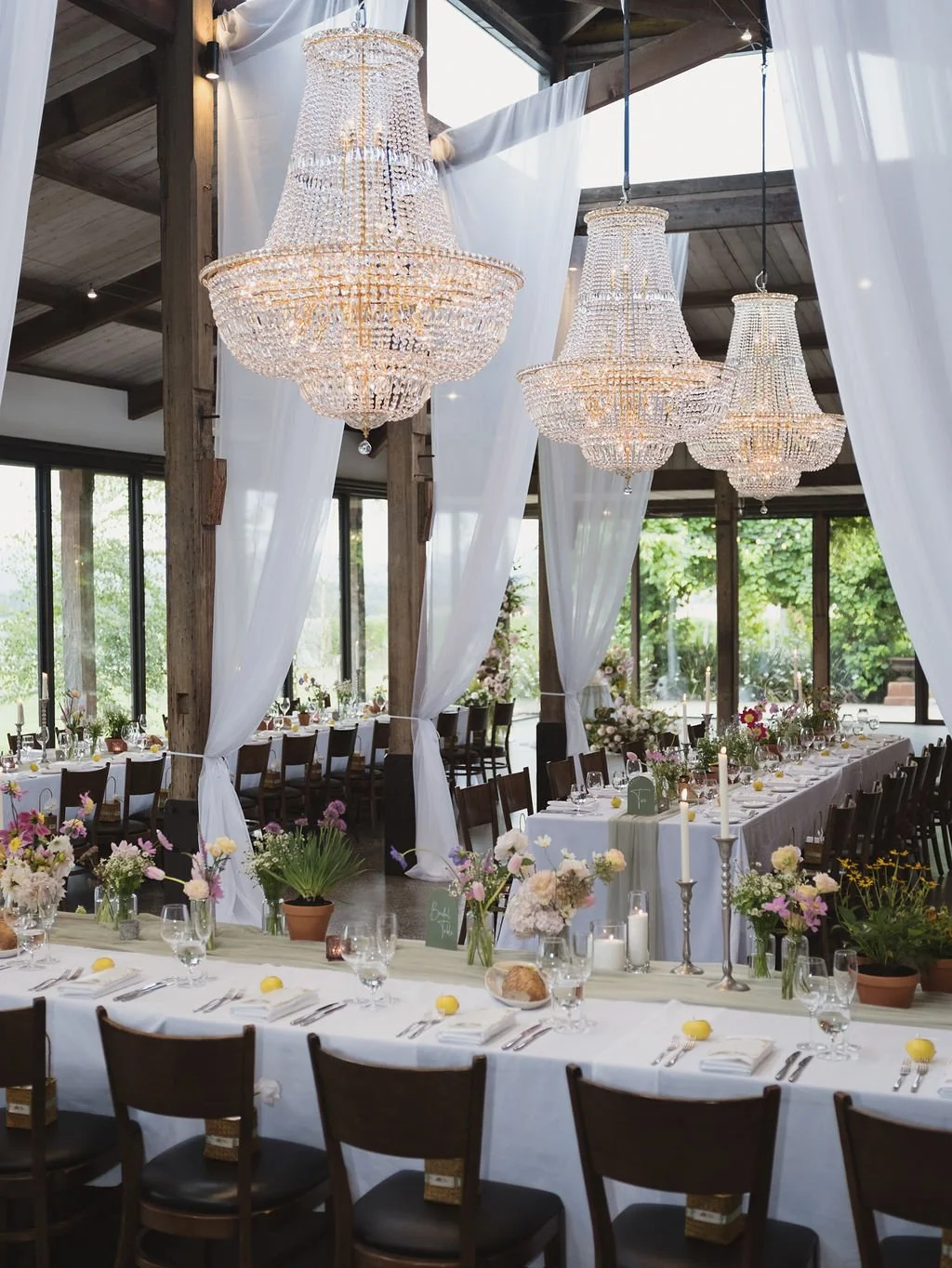 Elegant wedding reception setup with long banquet tables, white tablecloths, floral centerpieces, and six large crystal chandeliers hanging from the ceiling of a rustic venue with wooden beams and large windows.